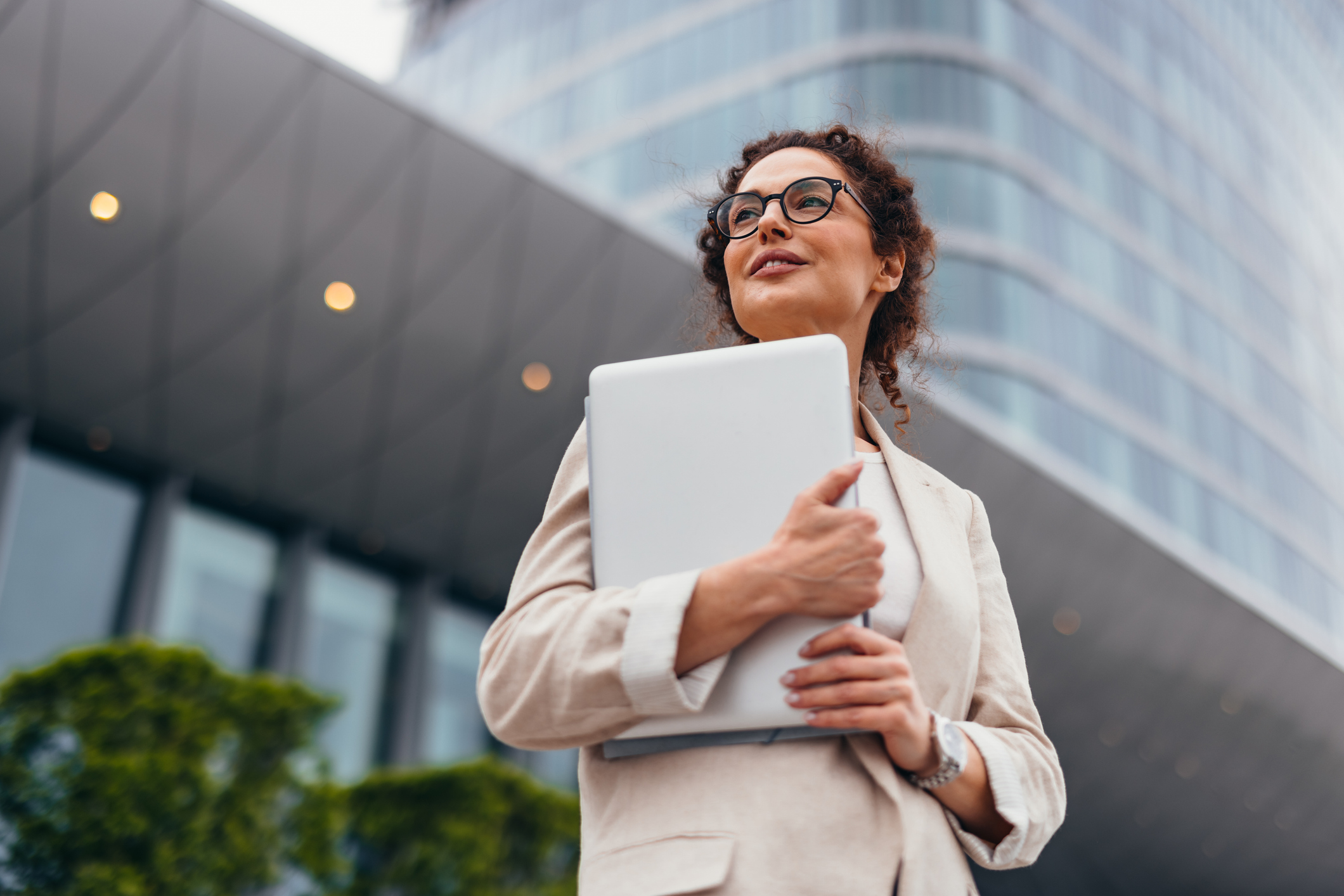 Smiling businesswoman in her 40s is holding a laptop in her arms while looking away and thinking about future projects in front of an office building