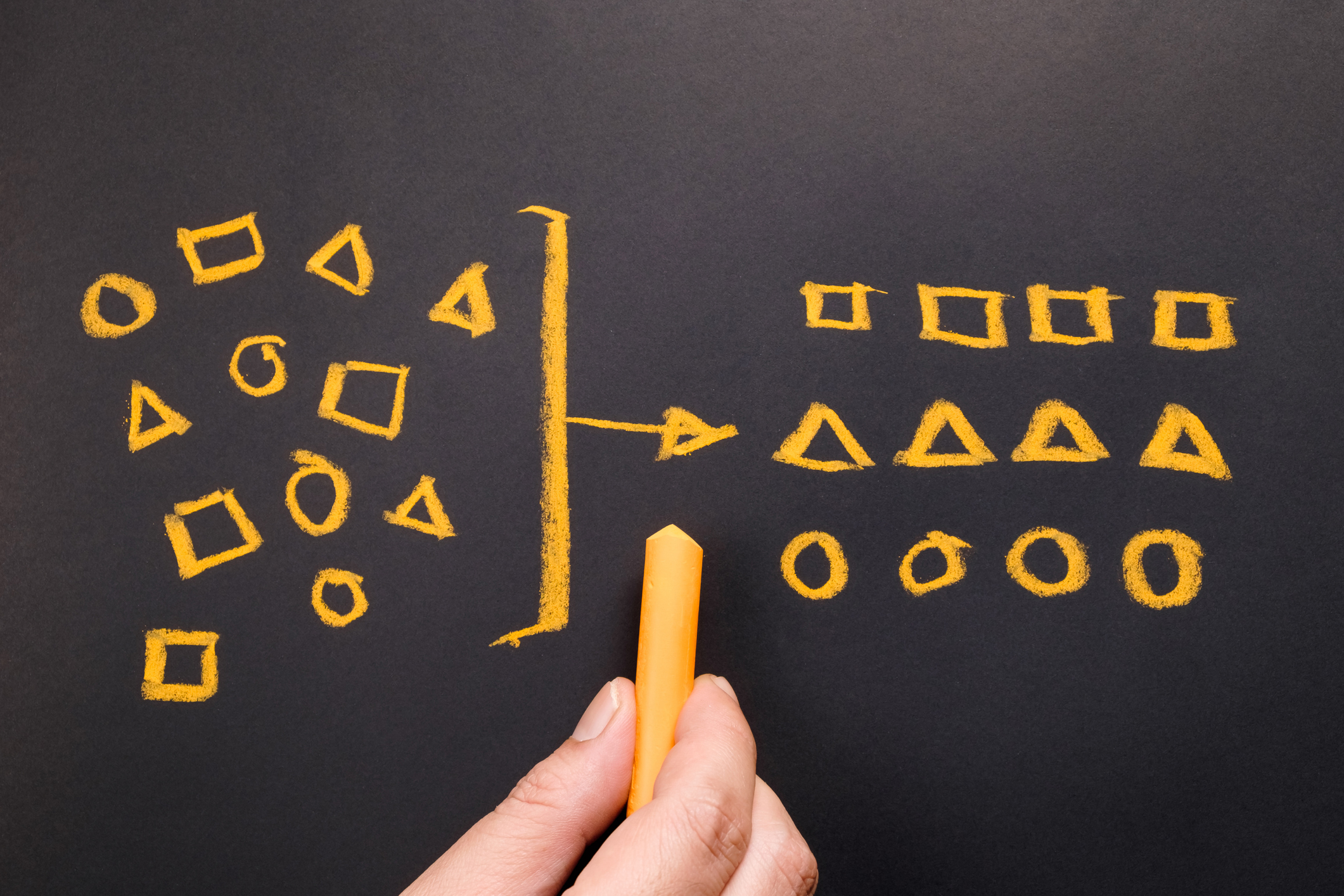 Hand drawing three sets of geometric shapes on a chalkboard, with an arrow arranging them a set of ordered groupings.