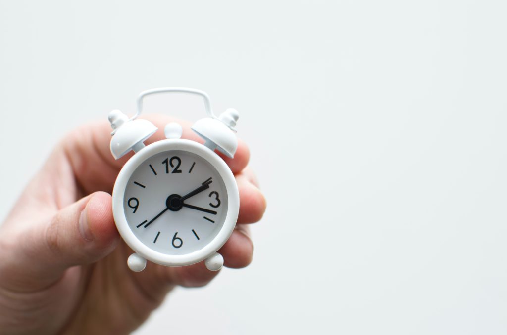 Hand holding a small, white alarm clock, with two bells on its top.