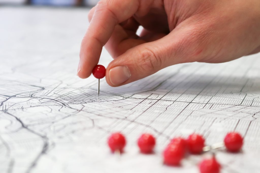 Close up image of a hand pressing a red push pin into a map. 