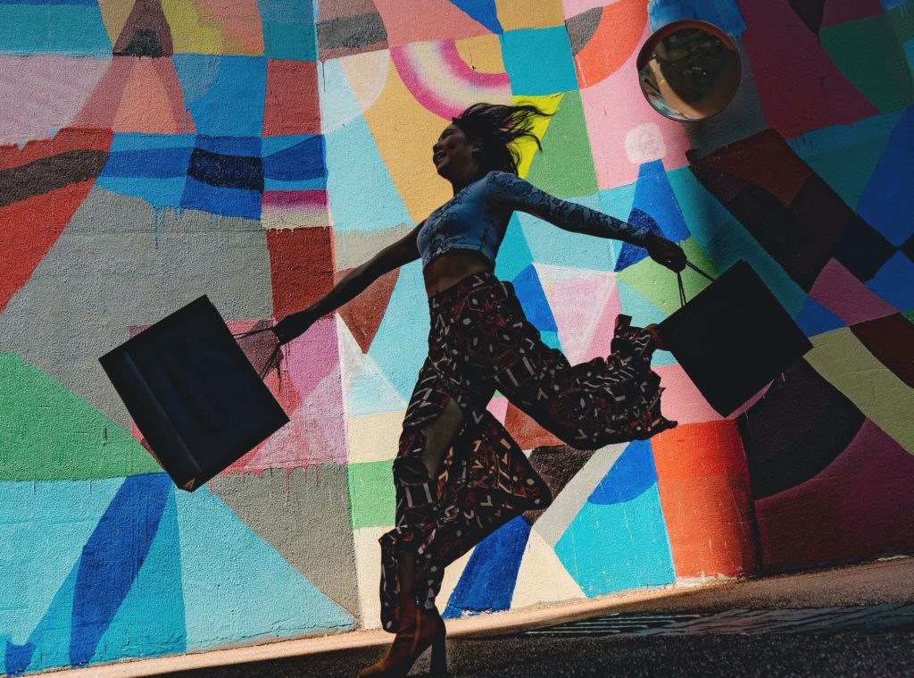 A smiling shopper, swinging shopping bags in both hands, walking past a colorful mural.