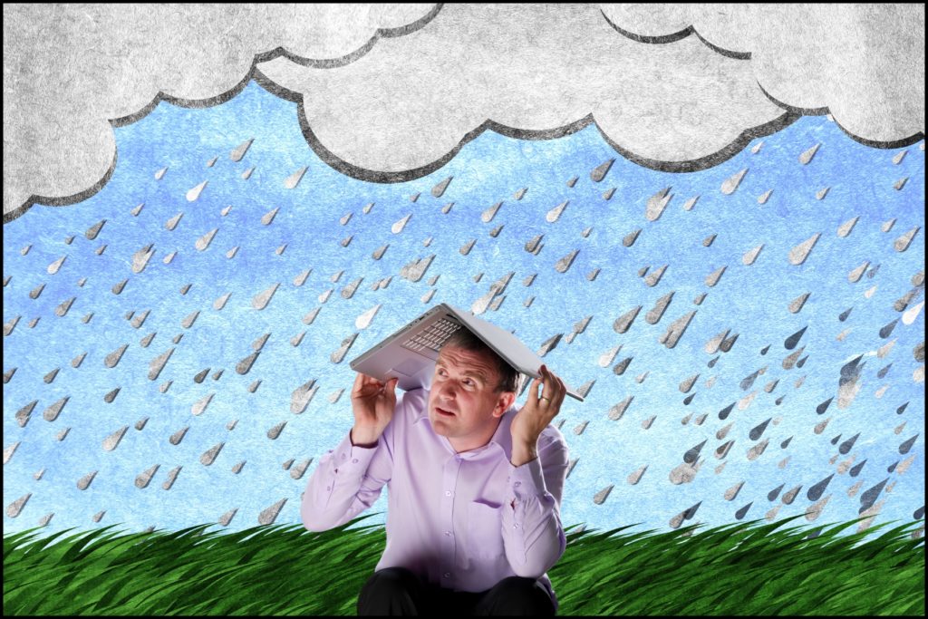 Tech worker holding a notebook computer over his head as a gray storm cloud rains overhead.