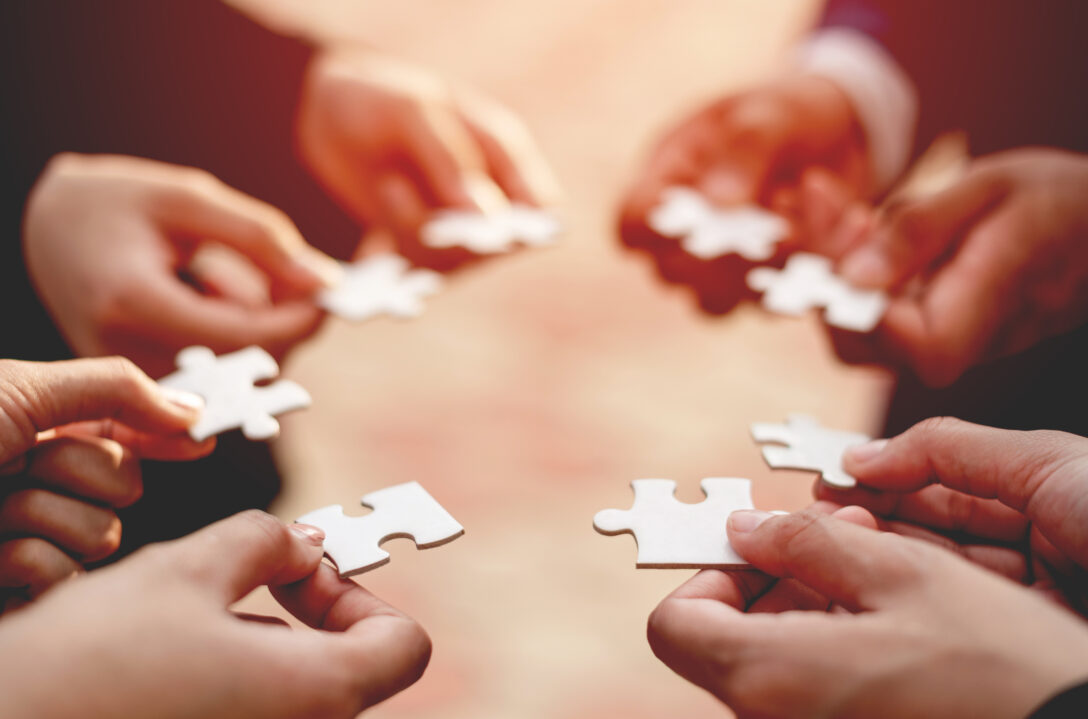 Close up of six hands forming a circle, each hand holding a white jigsaw puzzle piece.
