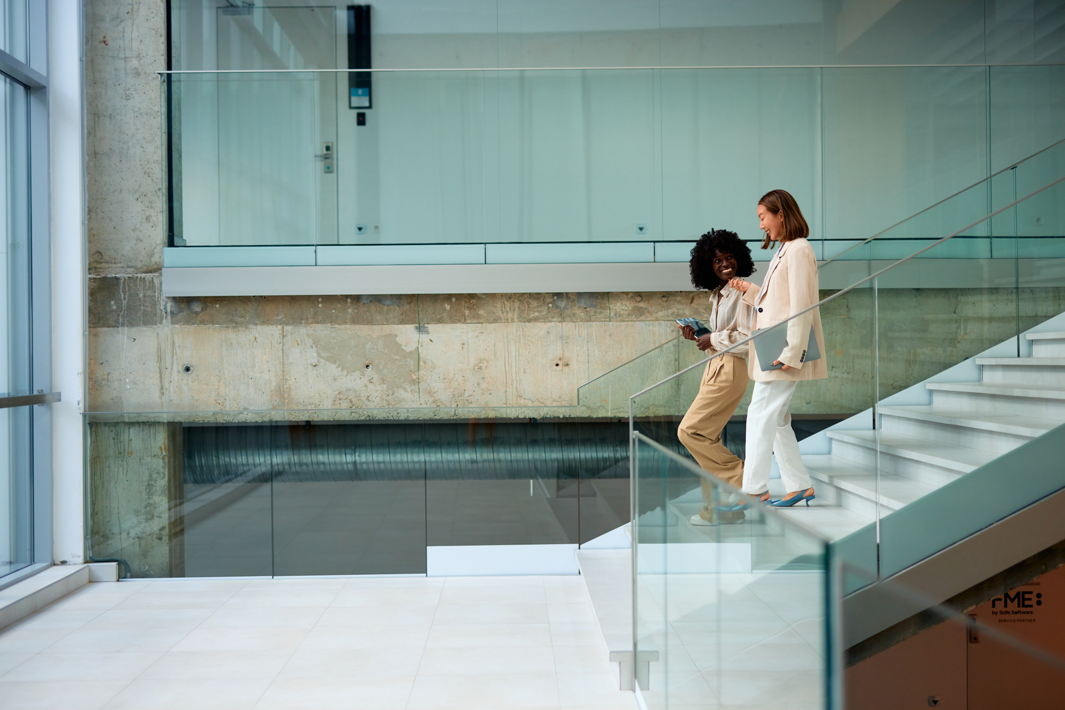 Two businesswomen climbing stairs in a modern office building, smiling and engaging in conversation while holding a laptop and a tablet, exemplifying teamwork and collaboration