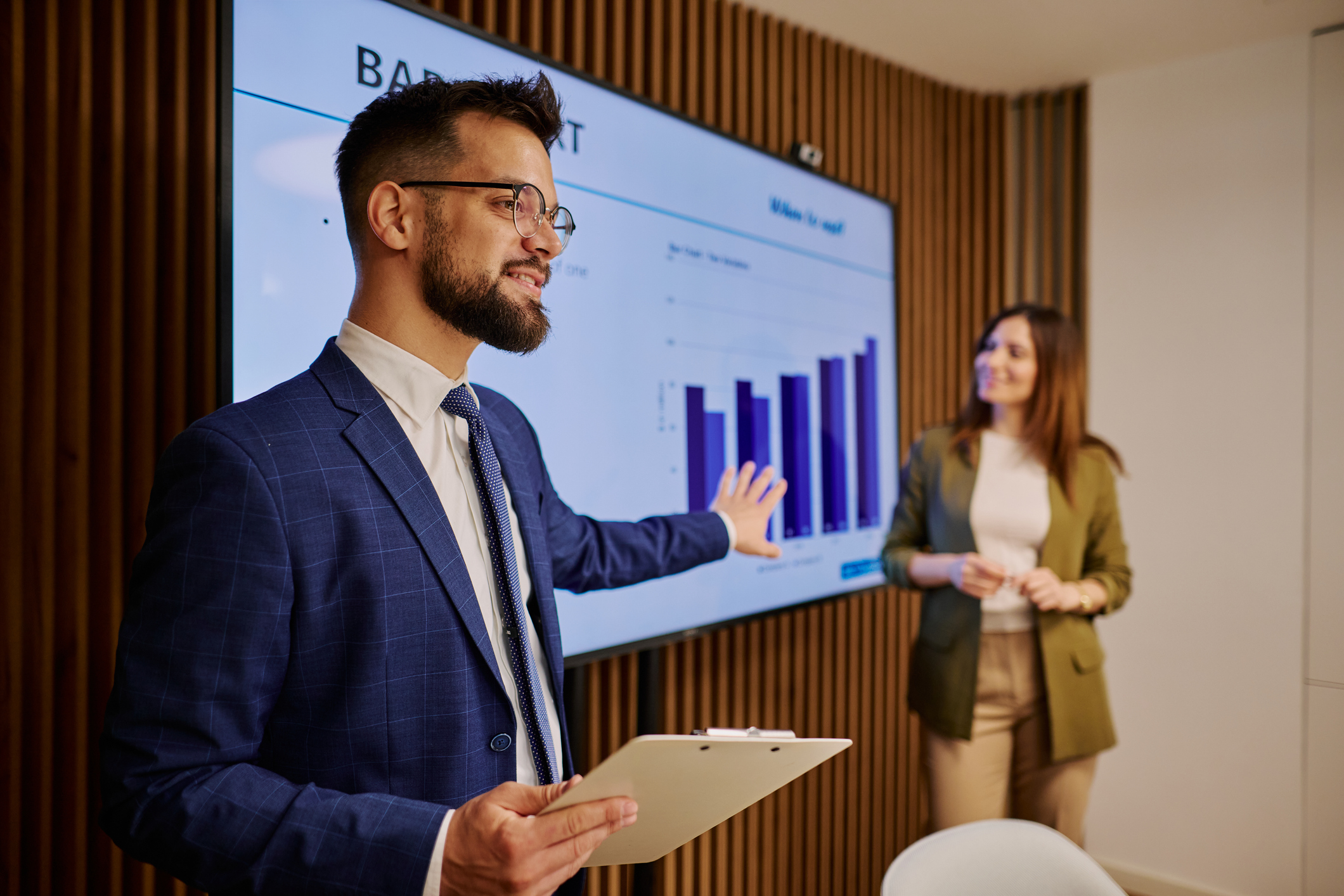Young businessman presenting positive bar chart growth on a large monitor, collaborating with a colleague in a sleek, modern meeting room, exemplifying professionalism and teamwork