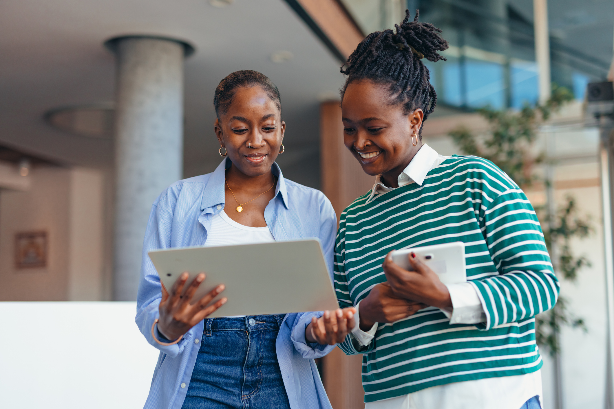 Two young businesswomen, smiling and engaged, are comparing data on a tablet and phone while standing in a sleek, modern office building, showcasing teamwork and collaboration
